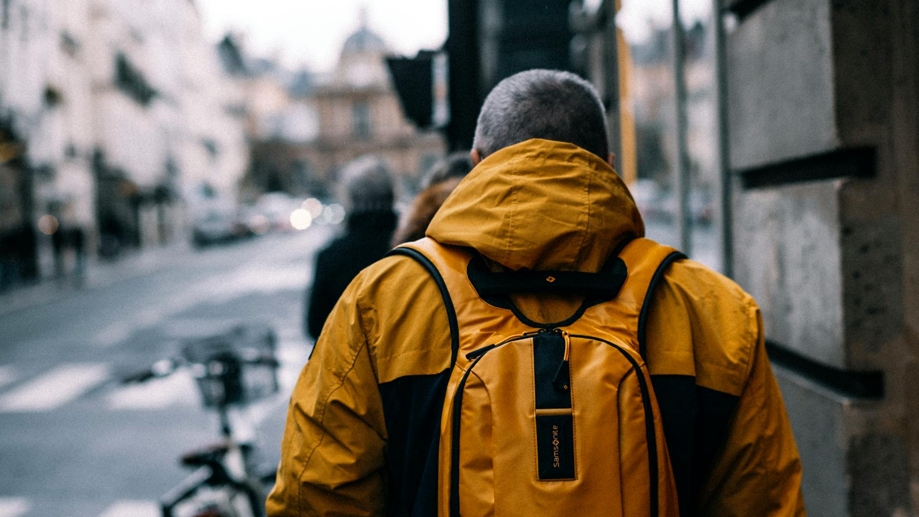 Back view of a man in a yellow jacket and backpack walking down a city street.