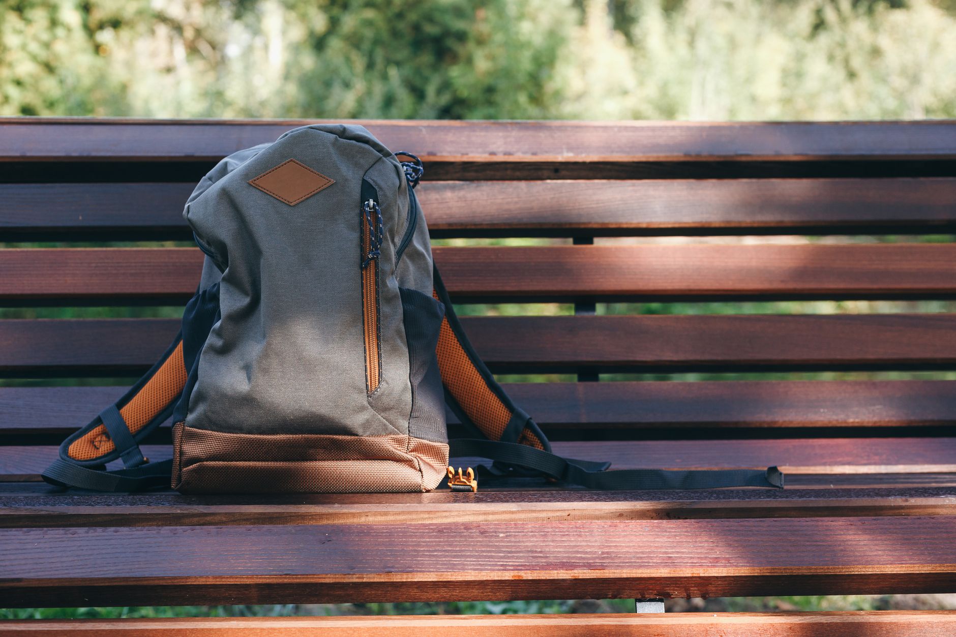 A brown and gray backpack resting on a wooden park bench in a sunny outdoor setting.