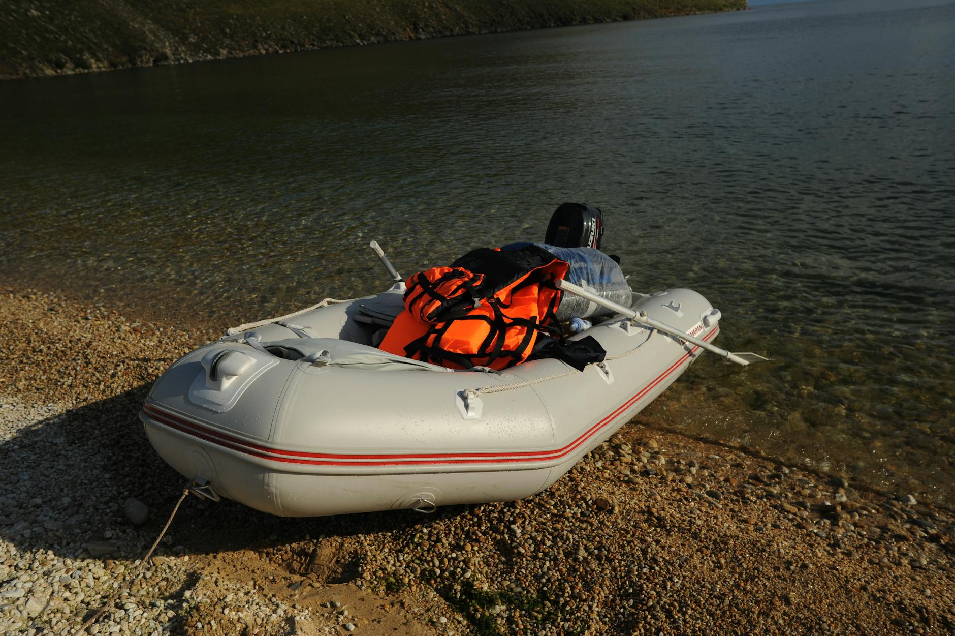 A white inflatable boat with an orange life vest rests on a peaceful rocky shore.